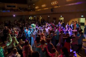 „Große Menschenmenge feiert gemeinsam in einem Saal, viele tanzen in einer langen Polonaise. Die Stimmung ist fröhlich, buntes Licht beleuchtet die Tanzenden, während andere am Rand sitzen oder zuschauen.“