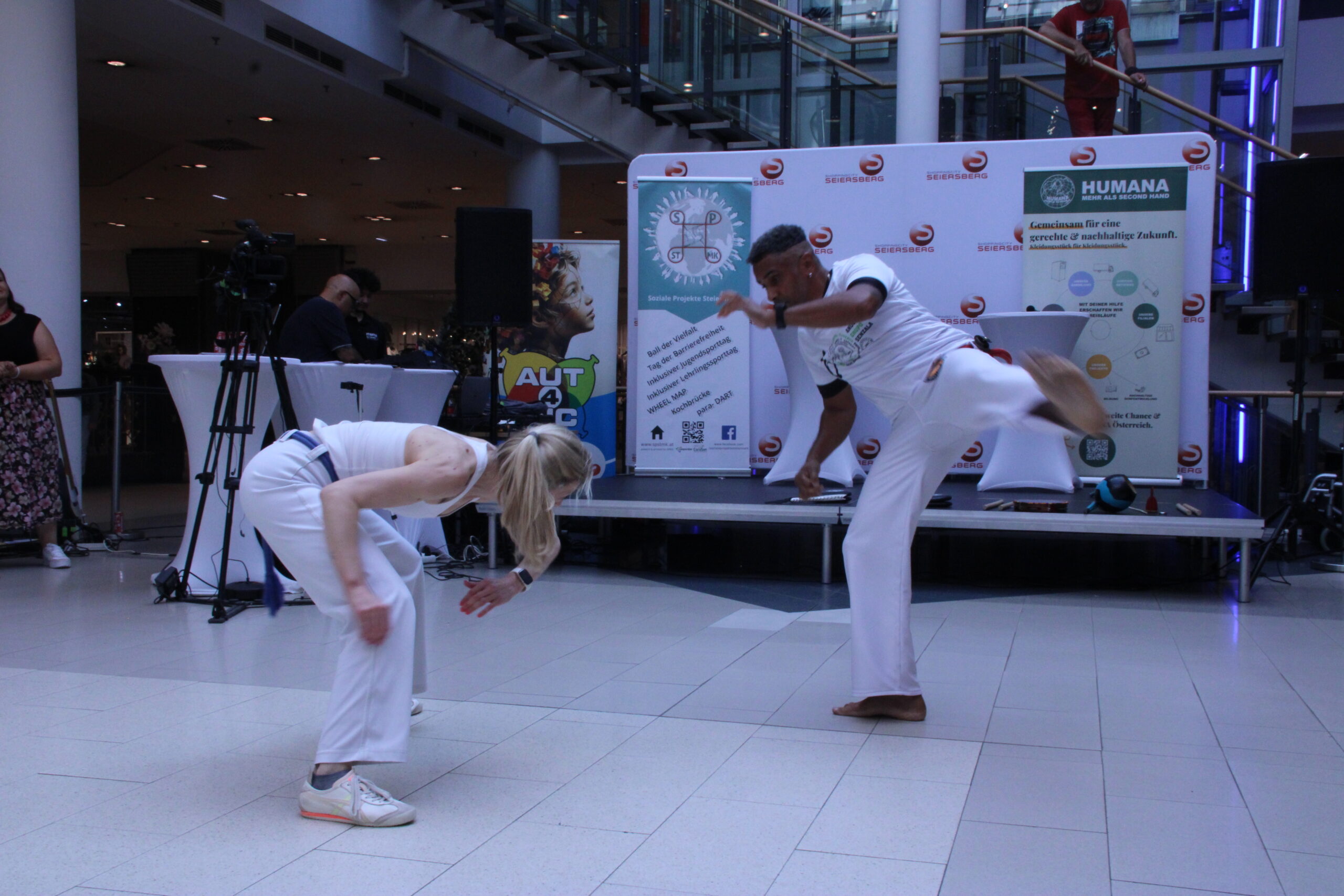 „Zwei Personen führen beim Inklusionstag in der Shopping City Seiersberg eine Capoeira-Tanz Vorführung auf. Eine Person weicht einer hohen Kickbewegung aus, während die andere einen Tritt ausführt.“