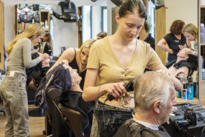 Szene in einem Friseursalon: Eine junge Friseurin schneidet einem älteren Mann die Haare, während andere Kundinnen behandelt werden. Soziale Projekte Steiermark könnten hier zur Förderung beitragen.
