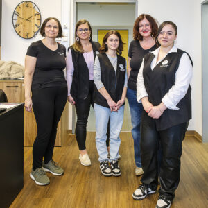 Fünf Frauen, stehen in einem hellen Büro mit Holzboden. Sie tragen schwarze Westen über ihren Outfits. Eine Uhr ist im Hintergrund sichtbar.