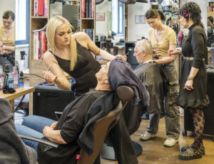 In einem Friseursalon: Blonde Friseurin färbt die Augenbrauen eines Kunden, während andere Friseure weitere Kunden bedienen. Regale mit "IGORA ROYAL" und "KOLESTON PERFECT" Produkten im Hintergrund.