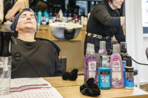 Eine Kundin entspannt mit Augenmaske im Friseursalon. Im Vordergrund stehen Guhl und John Frieda Haarpflegeprodukte wie Hyaluron Serum und Frizz Ease. "NEU GUHL HYALURON PFLEGE" ist sichtbar.