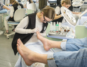 Eine Podologin behandelt die Füße eines Patienten in einer hellen Praxis. Sie trägt eine schwarze Weste und Handschuhe. Instrumente und Lotionen stehen bereit.