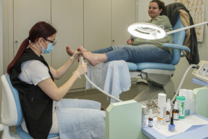 Eine Podologin mit rotem Haar und Maske behandelt die Füße einer Frau in Jeans unter einer Lampe. Diverse Instrumente und Flaschen stehen bereit. Fußpflege im Fokus.