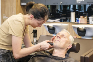 Friseur rasiert Bart eines älteren Mannes im Salon. Junge Friseurin mit Pferdeschwanz, die mit einem Trimmer arbeitet. Herrenfriseur im Friseursalon.