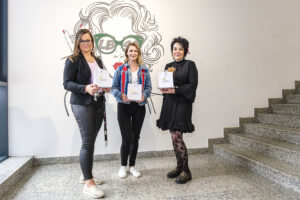 Drei Frauen posieren vor einer Wand mit dem Logo "LB" und einer Zeichnung einer Frau mit Brille. Sie halten weiße Geschenktüten und stehen vor einer Treppe.