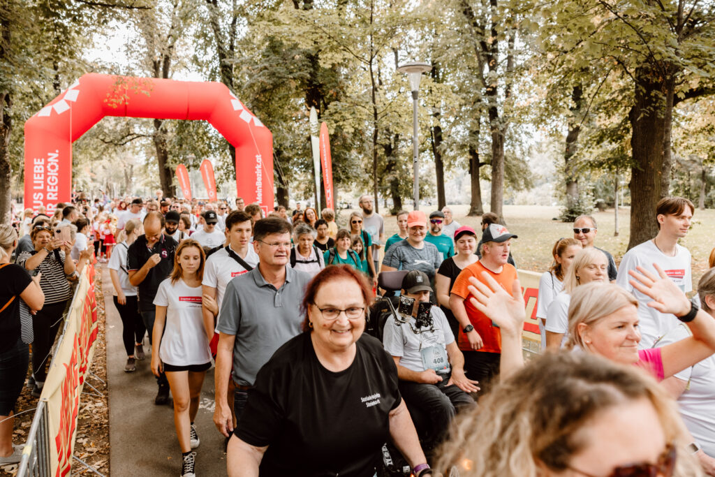 „Teilnehmerinnen und Teilnehmer eines inklusiven Laufs gehen gemeinsam durch einen Park unter einem roten Startbogen.“