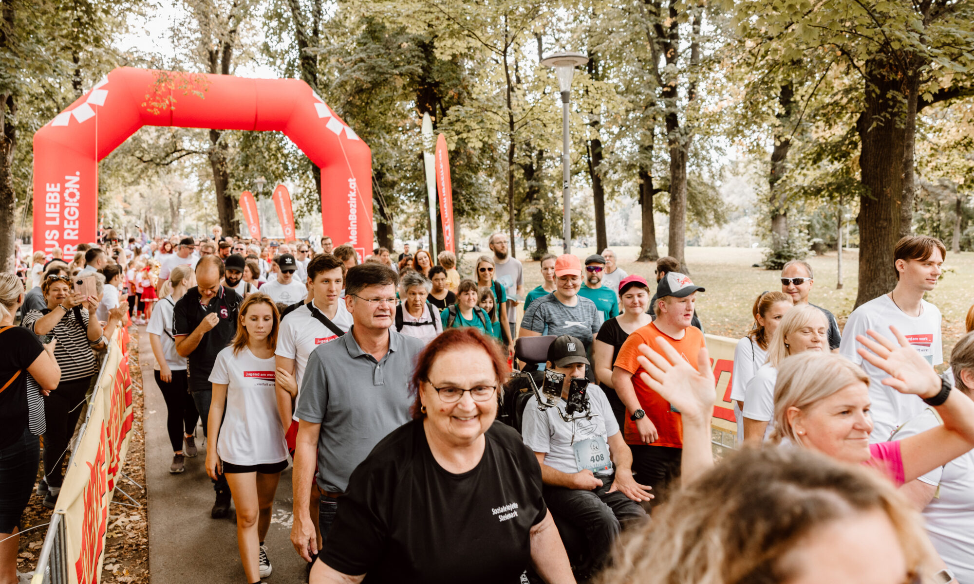 „Teilnehmerinnen und Teilnehmer eines inklusiven Laufs gehen gemeinsam durch einen Park unter einem roten Startbogen.“