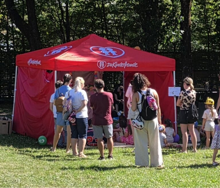 Rotes Zelt der "Die Kinderfreunde" mit Besuchern, die sich darunter auf dem bei einer Veranstaltung aufhalten. Kinder spielen im Hintergrund.