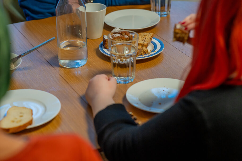 Menschen mit roten und grünen Haaren sitzen an einem Holztisch und essen Kuchen und trinken Wasser. Teller und Gläser sind auf dem Tisch verteilt.
© Bild von Achterbahn Steiermark