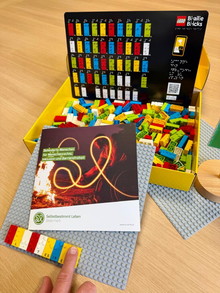 „Auf einem Tisch stehen LEGO Braille Bricks in einer gelben Box mit einer Übersichtstafel der Braillesteine. Daneben liegt eine graue LEGO Bauplatte mit einem Heft zum Thema Menschenrechte, Inklusion und Barrierefreiheit von Selbstbestimmt Leben Steiermark. Eine Hand zeigt auf eine mit LEGO Steinen gelegte Brailleschrift am unteren Rand der Bauplatte.“
© Bild von Selbstbestimmt Leben Steiermark