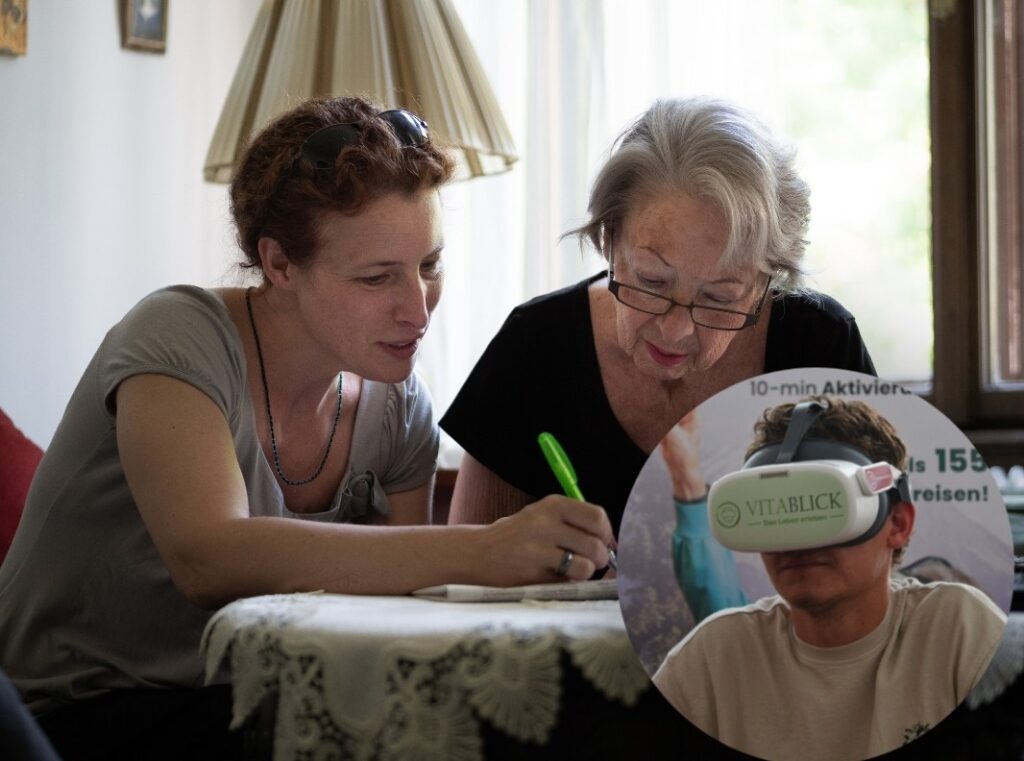 Zwei Frauen sitzen gemeinsam an einem Tisch und schreiben in einem Heft. Rechts unten ist ein rundes Bild eingeblendet, das einen Mann mit einer VR-Brille der Marke Vitablick zeigt.