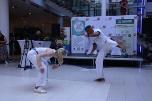 Zwei Capoeira-Kämpfer in weißen Anzügen führen eine akrobatische Darbietung auf, ein Mann tritt hoch, während eine Frau in einer tiefen Hocke ist. Im Hintergrund ist eine Bühne mit dem Logo "Programm Inklusionstag" zu sehen.