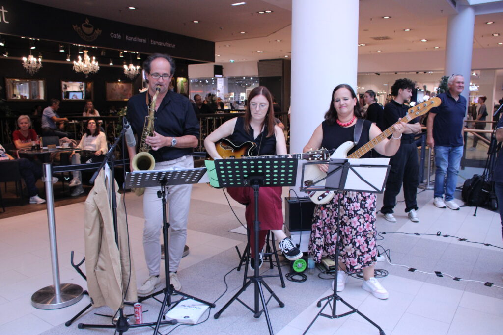 „Drei Musikerinnen und Musiker spielen beim Inklusionstag in der Shopping City Seiersberg gemeinsam Saxofon, Gitarre und E Bass. Das Publikum verfolgt den Auftritt im Einkaufszentrum.“