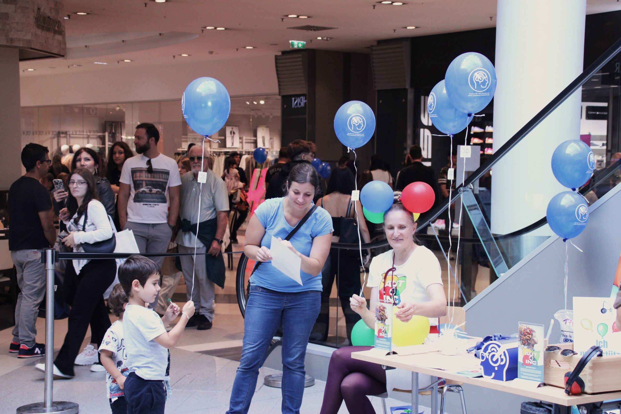 „Besucherinnen und Besucher informieren sich beim Inklusionstag in der Shopping City Seiersberg an einem Info Stand über Autismus. Eine Frau verteilt Informationsmaterial, während Kinder mit Luftballons am Stand spielen.“