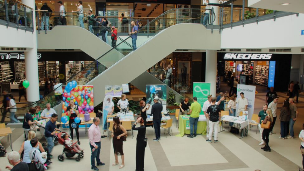 Blick in ein belebtes Einkaufszentrum mit mehreren Ebenen, Rolltreppen, Geschäften wie Levi's und Skechers, Ständen mit bunten Ballons und Infomaterial sowie vielen Menschen, die einkaufen und sich unterhalten.