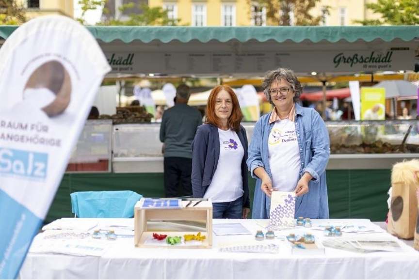Zwei Frauen stehen hinter einem Tisch mit Informationsmaterial, der für den Inklusionstag vorbereitet wurde. Im Hintergrund ist ein Marktstand zu sehen.